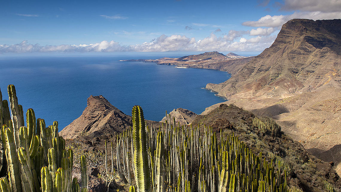 Steile Felsklippen an der Westküste Gran Canarias mit Blick auf den Atlantik. Im Vordergrund ragt eine Aussichtsplattform („Mirador del Balcón“) spektakulär über den Abgrund hinaus, auf der zwei Personen stehen. Darunter brechen die Wellen des Meeres an der felsigen Küste, während sich die zerklüfteten Klippen in die Ferne ziehen.